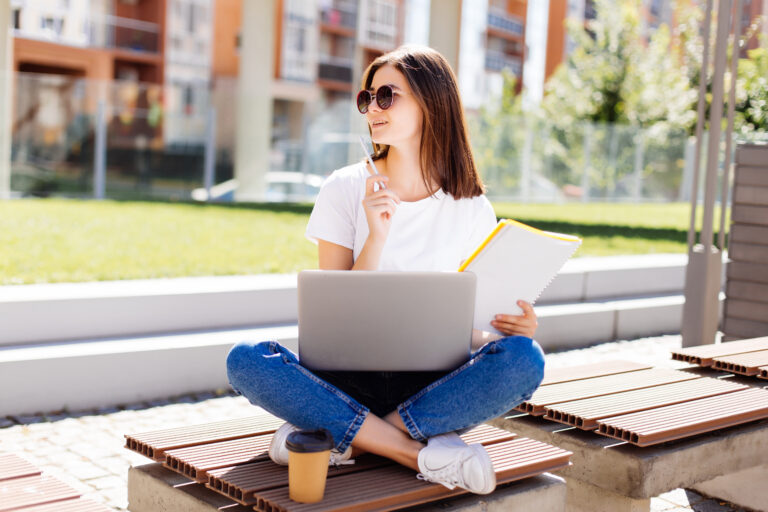 Confident and attractive woman sitting on bench in park and messaging in social media while writing in notebook during coffee break